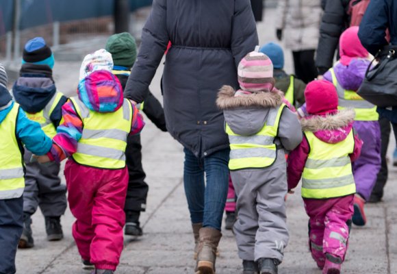 Barnehagebarn på tur i gule refleksvester, de har vinterdresser og lue, vi ser dem bakfra