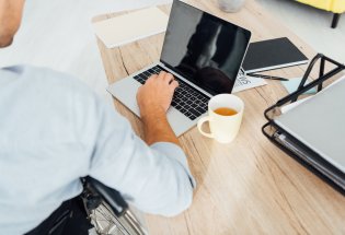 Mann i rullestol jobber på pc og drikker kaffe.