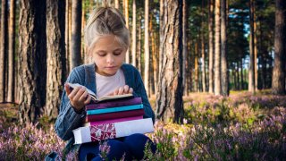 Jente som sitter i skogen og leser med en stabel bøker på fanget.